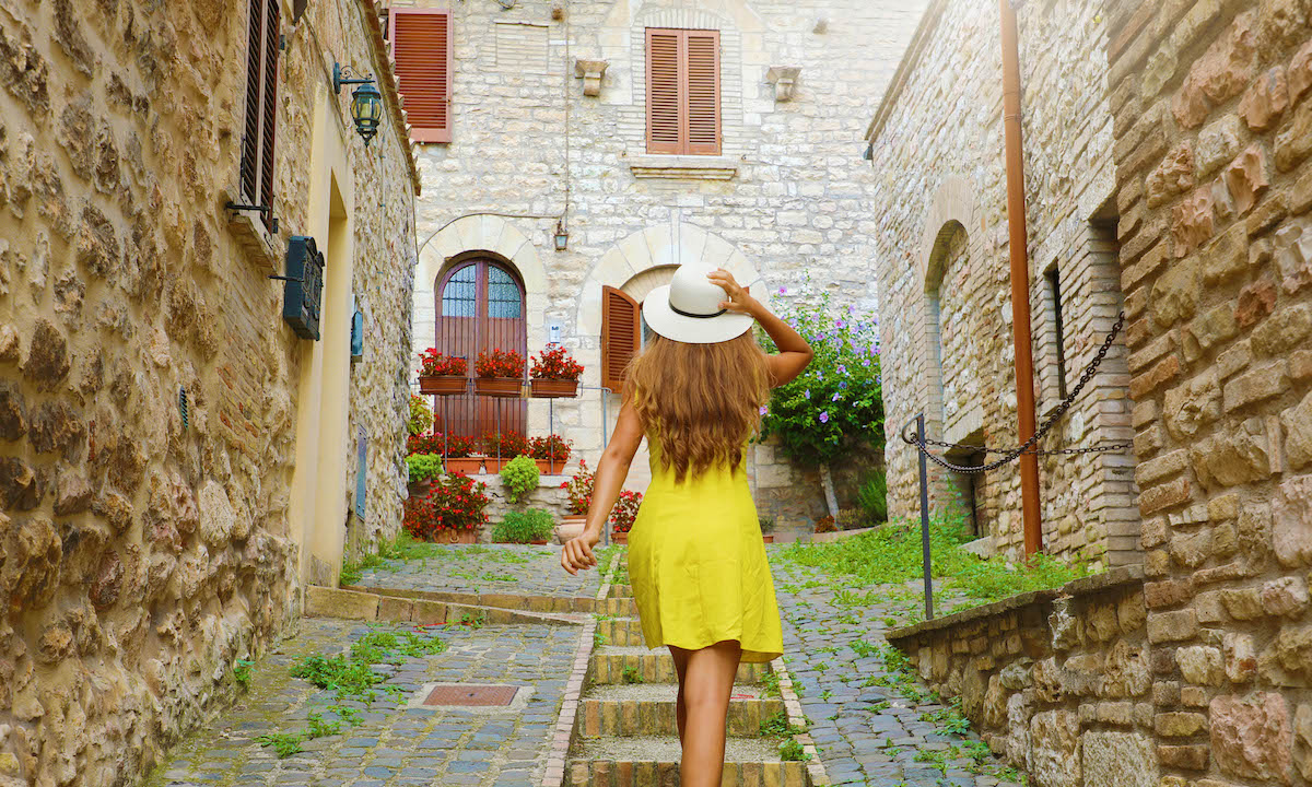 Beautiful curious young woman with yellow dress and hat goes upstairs in street in Assisi, Italy. Rear view of happy cheerful girl visiting central Italy.
