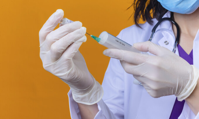 woman doctor wearing white coat with stethoscope in medical protective mask holding syringe and medicine vial vaccine bottle over isolated orange background